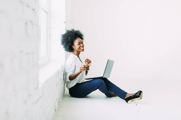 Young african american business woman using the laptop, while sitting on the floor near a big window in studio - Photo, Image