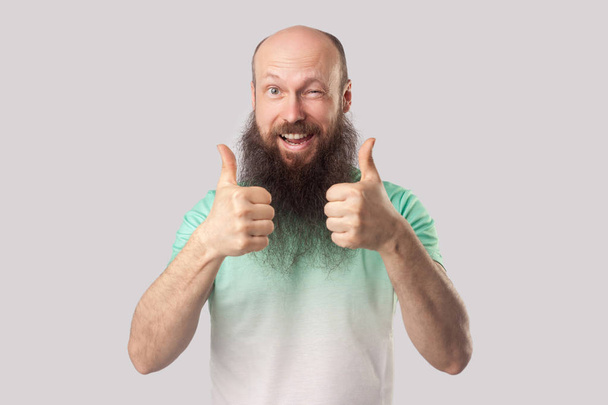 glad satisfied middle aged bald man with long beard in green t-shirt showing thumbs up and winking while looking at camera and toothy smile on grey background - Photo, Image