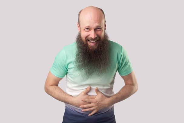 funny middle aged bald man with long beard in light green t-shirt holding his belly and laughing while looking at camera on grey background - Photo, Image