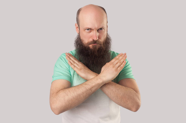 serious middle aged bald bearded man in green t-shirt showing cross sign and looking at camera on grey background - Φωτογραφία, εικόνα