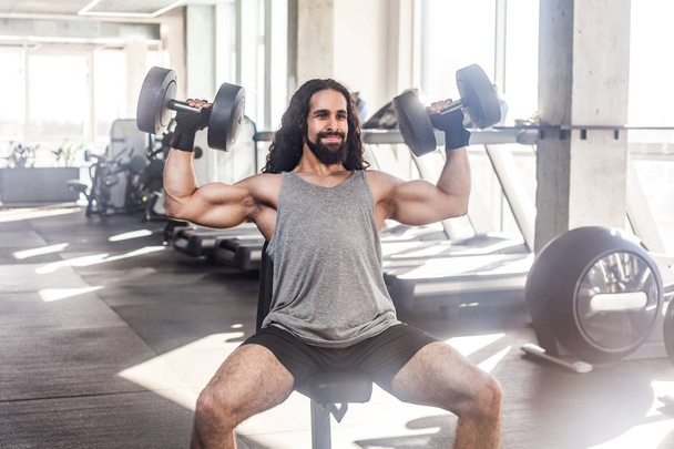 Fratze junger erwachsener muskulöser Athlet mit langen lockigen Haaren, der im Fitnessstudio trainiert, während er auf einem Gewichthebergerät sitzt und zwei Kurzhanteln mit erhobenen Armen hält, Übungen für schwingende Schultern  - Foto, Bild