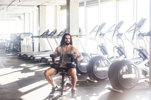 young adult muscular built handsome athlete with long curly hair sitting on weightlifting machine and holding two dumbbell on knees in gym  - Foto, immagini