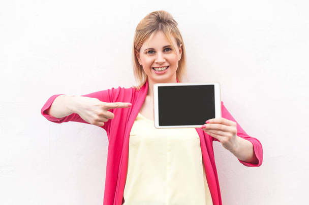 satisfied beautiful young businesswoman in pink blouse holding tablet pc and pointing finger to device and looking at camera with toothy smile isolated on white background - Фото, зображення