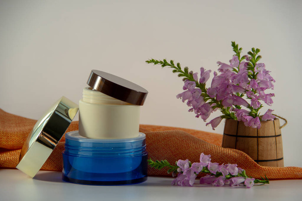 Deux paquets avec crème pour le visage, couvercles ouverts, tissu orange décoratif, vase en bois avec fleurs sauvages parfumées sur fond blanc
. - Photo, image