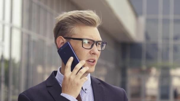 Young handsome Caucasian businessman in glasses talking on mobile phone in front of office building. Male executive manager in trendy clothes using smartphone outdoors. Modern technology concept - 映像、動画