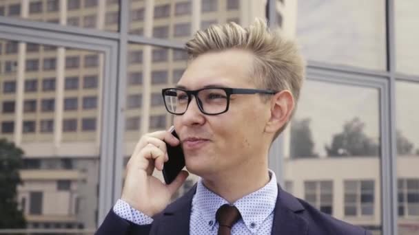 Fiatal csinos kaukázusi üzletember a szemüveg beszél mobiltelefonon előtt irodaépület. Férfi ügyvezető igazgató divatos ruhákat a szabadban okostelefon. Modern technológiai koncepció - Felvétel, videó