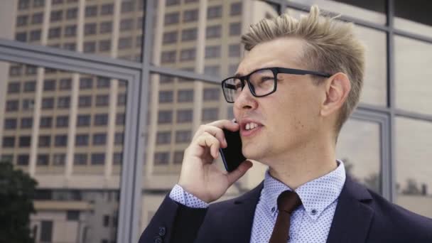 オフィスビルの前で携帯電話で話すメガネをかけた若いハンサムな白人ビジネスマン。屋外でスマートフォンを使用してトレンディな服を着た男性エグゼクティブマネージャー。現代技術の概念 - 映像、動画