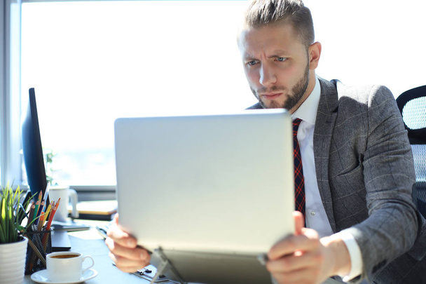 Good looking young man in full suit holding laptop while sitting in the office. - Foto, Bild