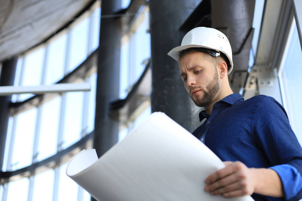 Confident young architect in navy shirt and hardhat holding a blueprint and looking at it. - Photo, Image