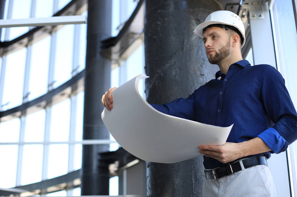 Confident young architect in navy shirt and hardhat holding a blueprint and looking at it. - Photo, Image