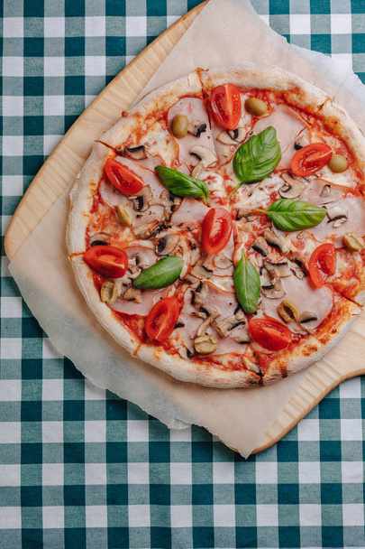 Neapolitan pizza with mushrooms, cheese, arugula, basil, tomatoes sprinkled with cheese on a wooden board on a tablecloth in a cell with a place for the text - Zdjęcie, obraz