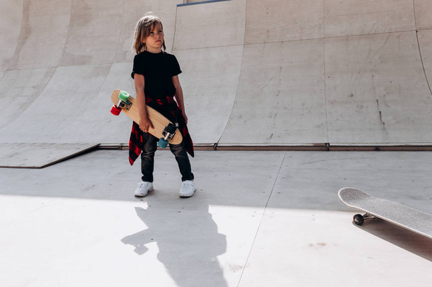 The boy dressed in the casual clothes with skateboard in his hand stands in a skate park next to the slide at the sunny day outside - Foto, imagen