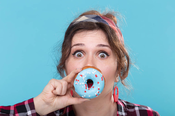 Nahaufnahme Porträt einer lustigen jungen schönen Frau mit Donut in der Nähe des Mundes, die auf blauem Hintergrund posiert. Konzept der schädlichen und kalorienreichen Desserts. - Foto, Bild