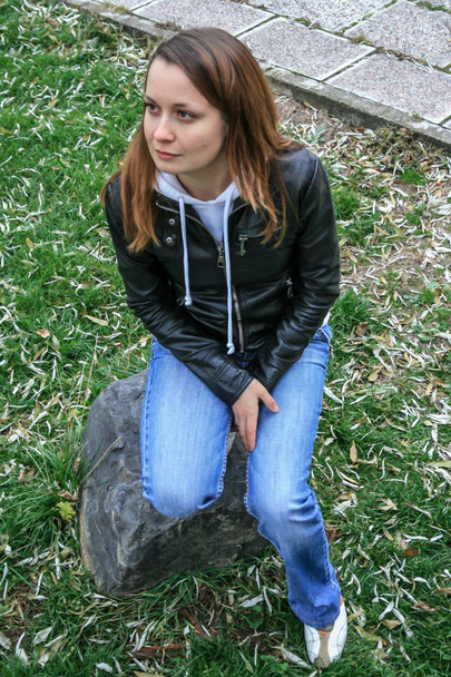 2010.11.19, Maloyaroslavets, Russia. A young woman wearing black jacket and blue jeans sitting on the stone in the park. - Фото, зображення