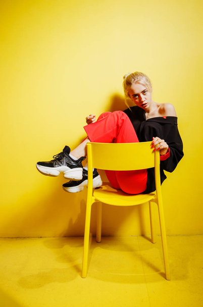 Young fashion girl blogger dressed in red trousers and black jacke possing sitting on the stool in the room with yellow walls and furniture - Фото, зображення
