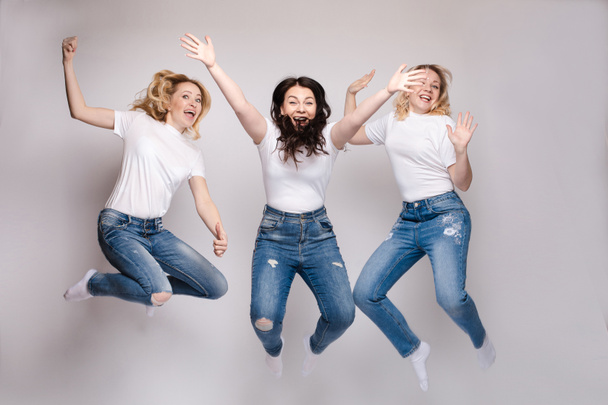 Three friends in casual clothes dancing and jumping. Different, but beautiful ladies in jeans and white T-shirts getting fun and smiling. Happy young blonde and brunette women spending time together. - Valokuva, kuva
