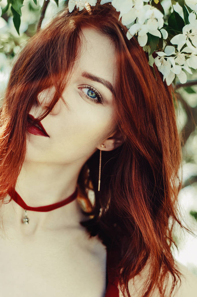 Kaunis nuori nainen, joka nauttii tuoksusta kukkivassa kevätpuutarhassa. Punaiset hiukset, siniset silmät ja punainen mekko, pitää kukkia käsissään
. - Valokuva, kuva