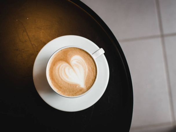 Uma xícara de cappuccino com arte latte na mesa preta, xícara de cerâmica branca, vista superior. Café cultura
. - Foto, Imagem