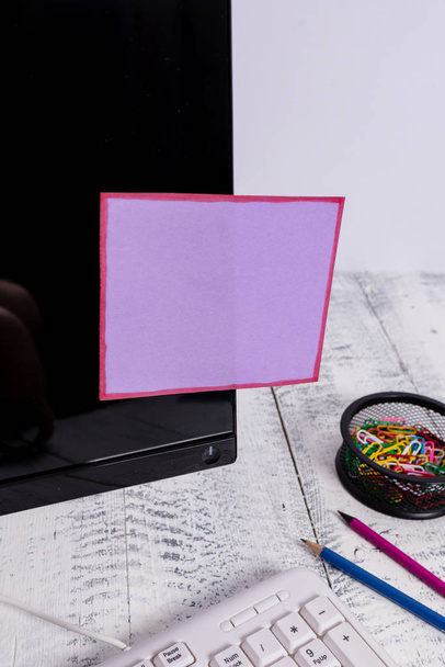 Notacja papier trzymać się czarny ekran komputera w pobliżu białej klawiatury i nieruchomy nad drewnianym stole. Monitor PC umieszczony w pobliżu urządzenia do pisania z purpurową taśmą samoprzylepna na nim. - Zdjęcie, obraz