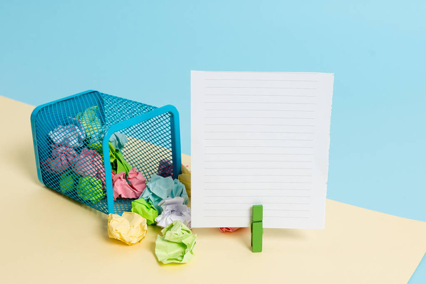 Little tipped trash bin full of crumpled colored paper and a clothespin hanging a colorful paper note in a yellow and blue background. Office supplies and empty reminder. - Photo, Image