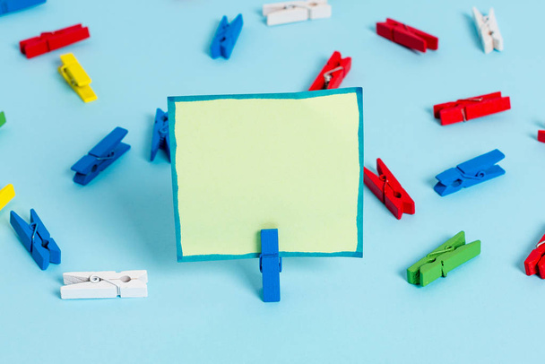 Colored clothespin placed around the empty rectangle square shaped reminder note in the centre of a blue background. Office supplies, blank paper pinned by a fastener. - Photo, Image