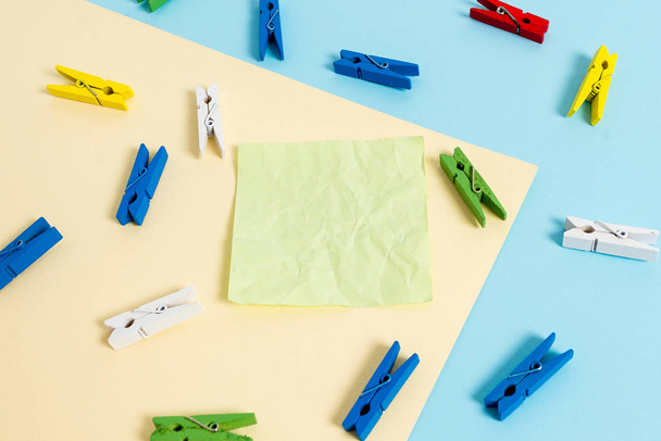Colored clothespin placed around the empty rectangle shaped reminder note in the centre of a blue background and yellow square. Office supplies, blank paper pinned by a fastener. - Photo, Image