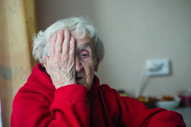 Stara kobieta w czerwonej kurtce pokrywa jedno oko ręką.  - Zdjęcie, obraz