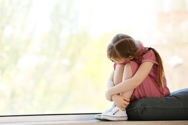 Θλιμμένο κοριτσάκι με αυτιστική διαταραχή που κάθεται στο περβάζι του παραθύρου στο σπίτι - Φωτογραφία, εικόνα