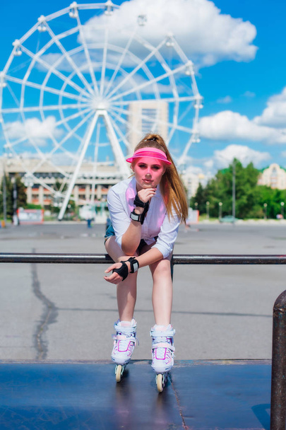 Portrait of an emotional girl in a pink cap visor wearing protective gloves and rollerblades sitting on the background of ferris wheel. - Fotografie, Obrázek