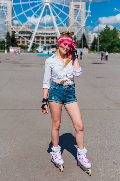 Portrait of an emotional girl in a pink cap visor and protective gloves for rollerblades and skateboarding riding on rollerblades on the road. - Foto, afbeelding