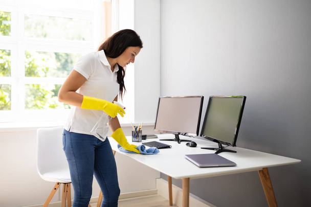 Side View Of Female Janitor Cleaning Desk With Blue Napkin In Office - Foto, afbeelding