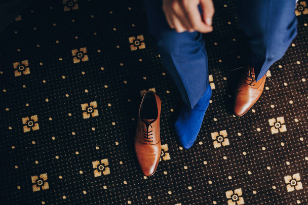 Stylish groom in blue suit putting on brown shoes near window in - Photo, image