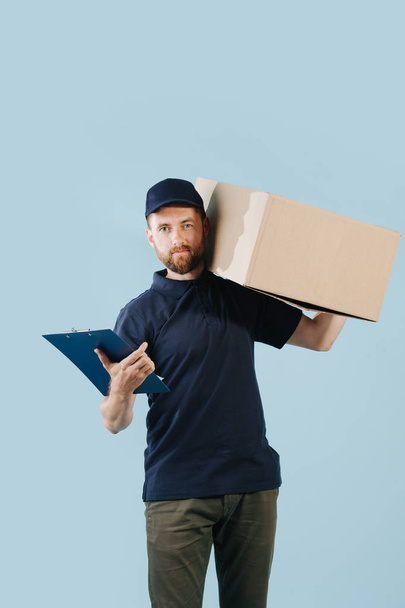 Delivery man in uniform is holding big cardboard box on his shoulder - Zdjęcie, obraz