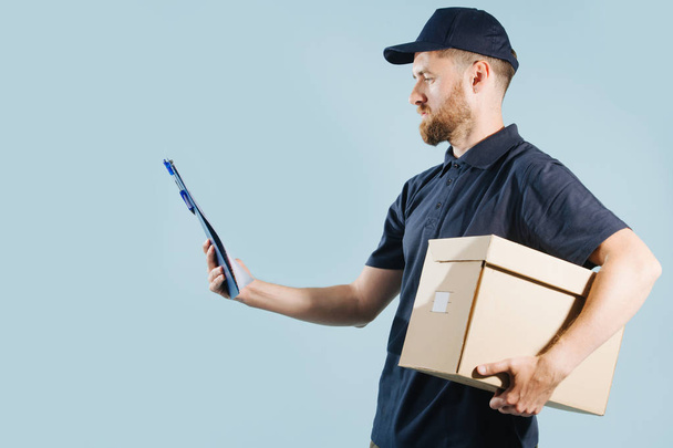 Delivery man in uniform is checking his orders list while holding box under arm - Zdjęcie, obraz