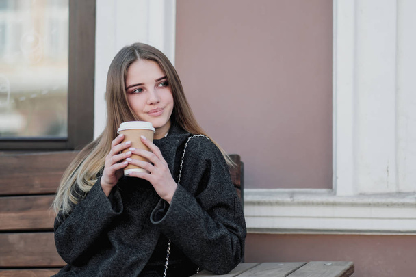 コートを着た若い美しいミレニアムの女の子は、コーヒーの紙コップとアウトドールベンチに座っています。秋または春の日、涼しい天気. - 写真・画像