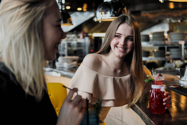 Две молодые умные красивые девушки в платьях сидят в баре. Пейте красные и синие коктейли из стаканов с трубочками
. - Фото, изображение
