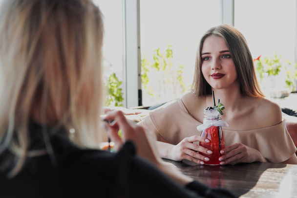 Two young smart beautiful girls in dresses sit at a restaurant table opposite a large window. Drink red and blue cocktails from glasses with tubules. - Foto, immagini