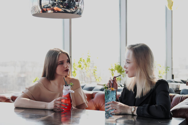 Two young smart beautiful girls in dresses sit at a restaurant table opposite a large window. Drink red and blue cocktails from glasses with tubules. - Fotó, kép