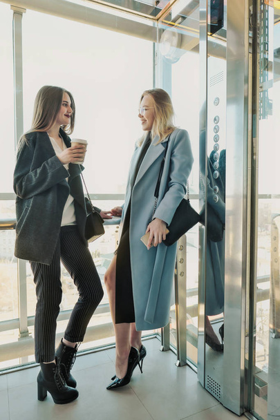 Two beautiful girls, a blonde and a brunette, are wearing a coat in a glass elevator with a window. Shopping center or office in the city. - Foto, afbeelding