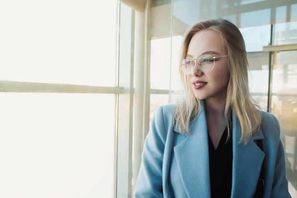 Portrait of a young beautiful girl with glasses and a blue coat in the elevator at the glass. - Foto, afbeelding