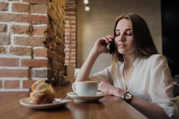 Młoda piękna brunetka dziewczyna w kawiarni w stylu loftu siedzi przy stole przy oknie, pije kawę i używa smartfona, połączeń, robi zdjęcia. - Zdjęcie, obraz