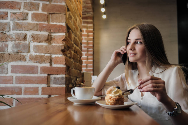 A young beautiful brunette girl in a loft-style cafe sits at a table by the window, drinks coffee and uses a smartphone, calls, takes pictures. - Foto, Bild