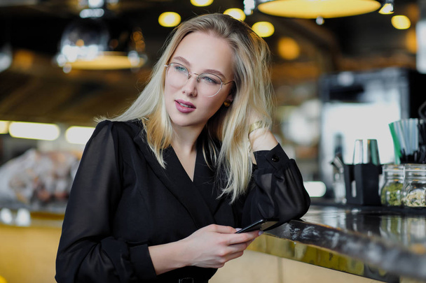 A young beautiful blond woman with glasses sits at the bar in a yellow interior. Drinks alcoholic cocktail with a straw and green mint. - Zdjęcie, obraz