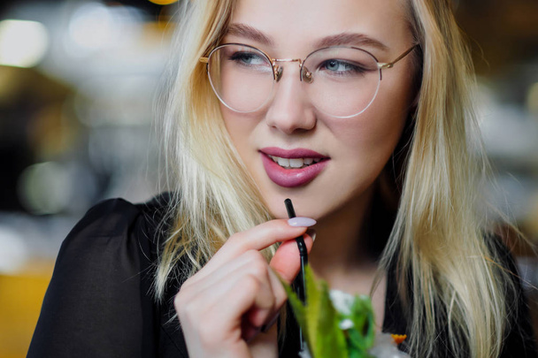A young beautiful blond woman with glasses sits at the bar in a yellow interior. Drinks alcoholic cocktail with a straw and green mint. - Фото, зображення