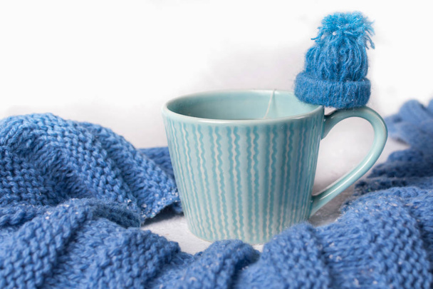 Winter concept. Blue white color . A cup of hot tea flavored with a knitted scarf and little hat on white backgroung. Cold color. - Foto, Imagem