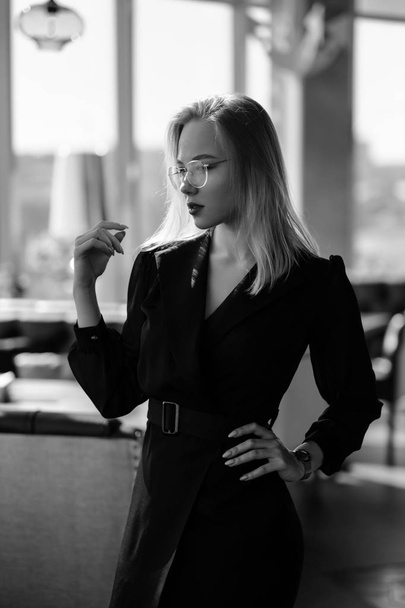 A young beautiful blond woman with glasses sits at the bar in a yellow interior. Drinks alcoholic cocktail with a straw and green mint. - Foto, afbeelding