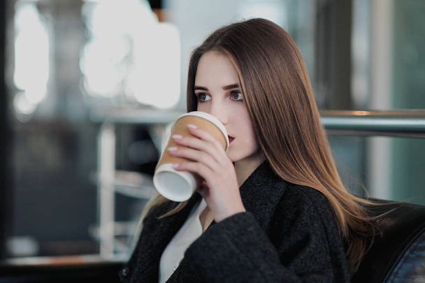 A young beautiful brunette girl in a gray coat is sitting on a bench waiting. Drinking coffee and holding a paper cup with a drink. - Foto, imagen