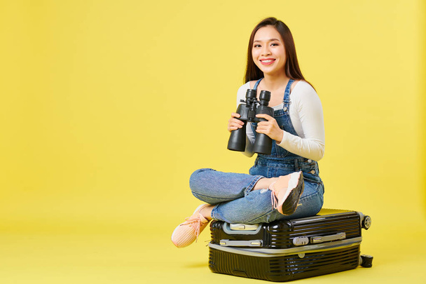Ευτυχισμένο όμορφο νεαρό ασιατικό γυναίκα σε ντένιμ συνολικά κάθεται σε βαλίτσα με κιάλια και κοιτάζοντας την κάμερα - Φωτογραφία, εικόνα