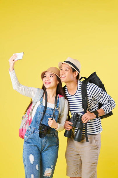 Cheerful young Vietnamese tourists with digital camera and binoculars posing for selfie on smartphone, isolated on yellow - Foto, immagini