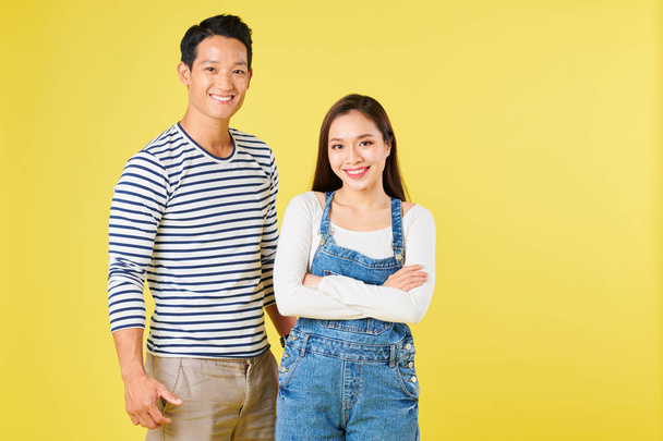Portrait of cheerful young confident Vietnamese couple in casual clothes standing against yellow studio background - Photo, Image